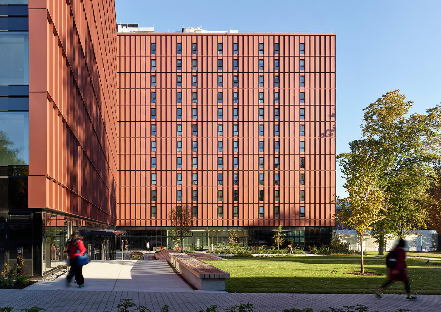 Exterior view of a building showcasing red panels and glass elements, with a landscaped area and people walking in the foreground.