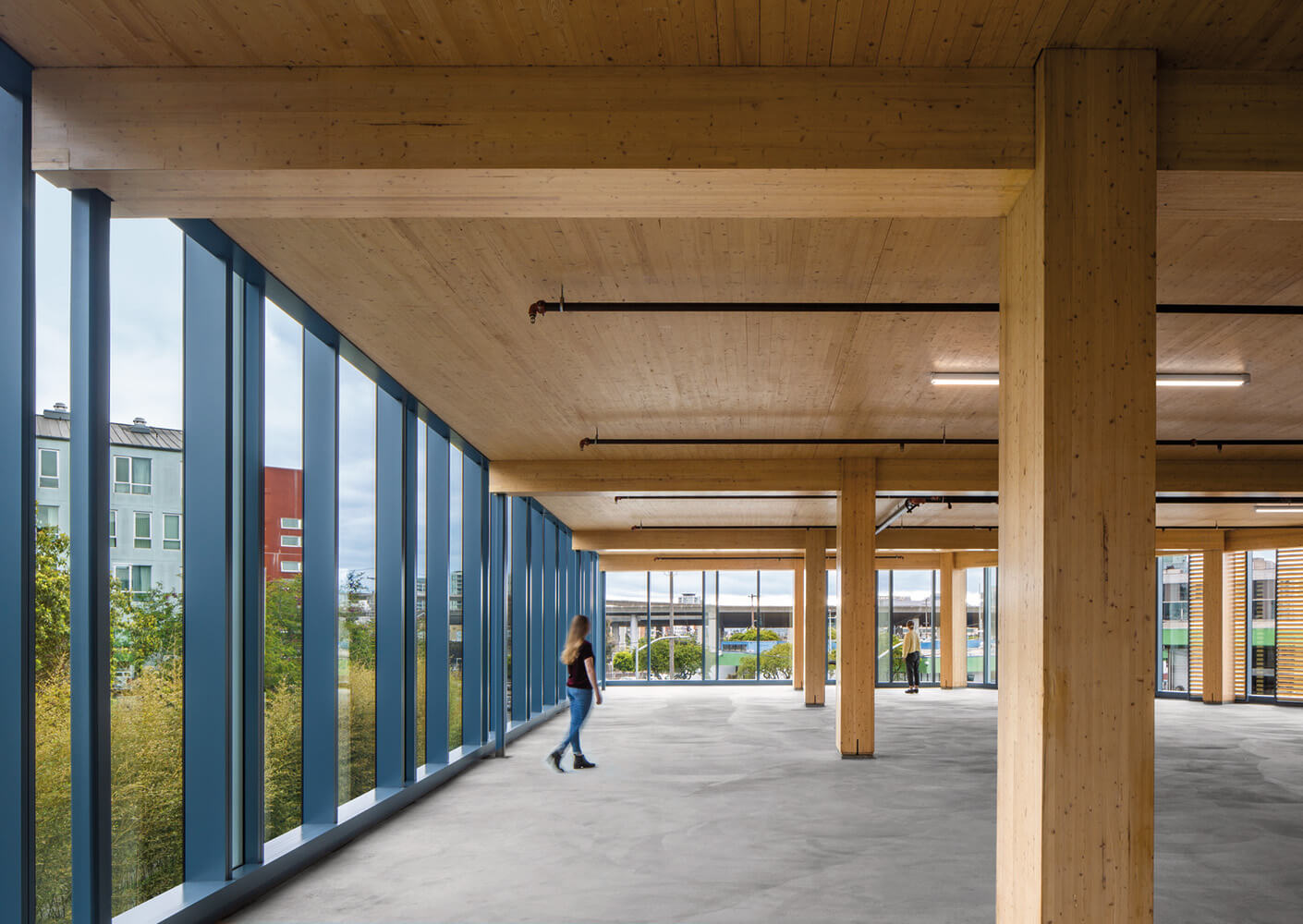 Spacious interior featuring wooden beams and large windows, with people walking and views of greenery and nearby buildings.