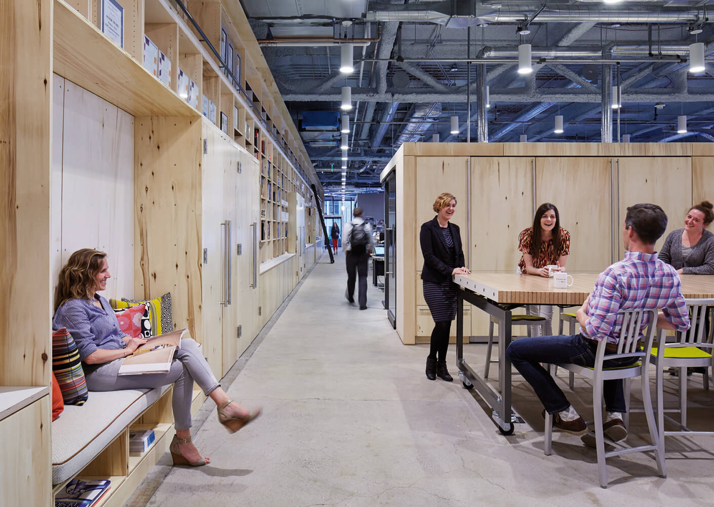 Casual office space featuring a collaborative area with a large table, seating, and wooden accents. Employees engage in discussions and reading.