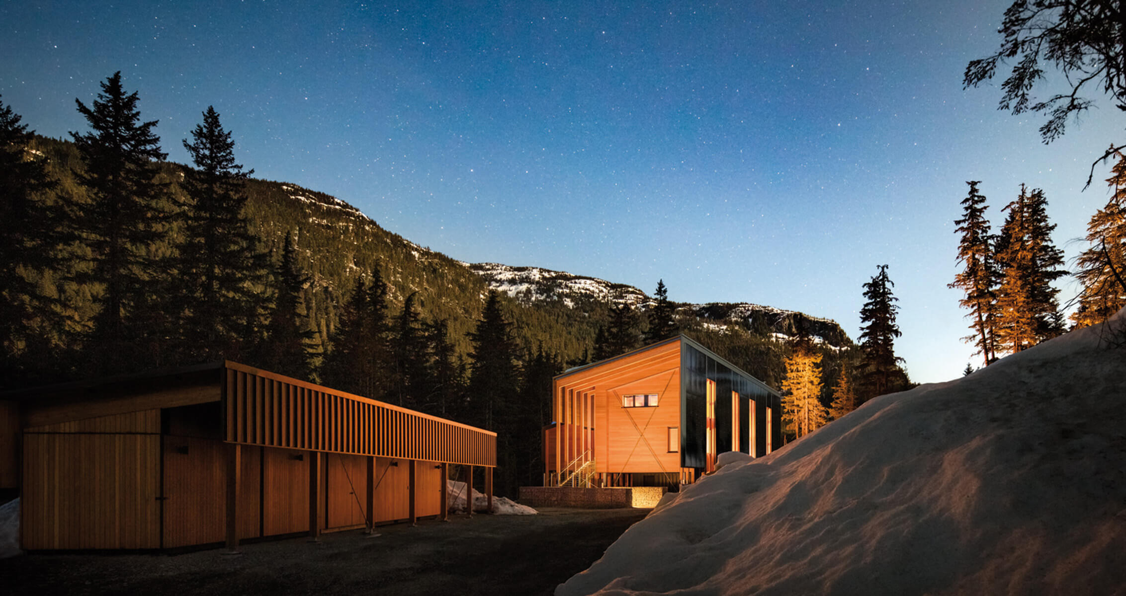 House surrounded by snow and trees in a mountainous area, illuminated under a starry night sky. Two structures visible from the side angle.