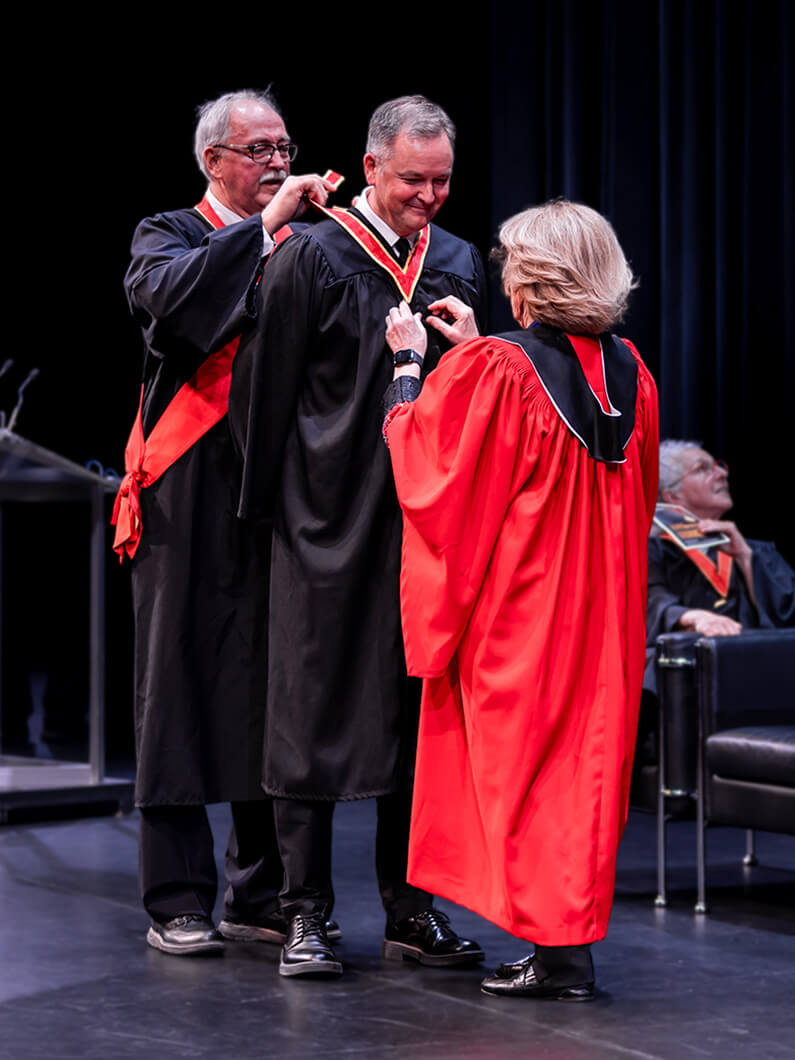 Three individuals on stage at a graduation ceremony, with one person placing a medal around another's neck. Black and red academic regalia is visible.