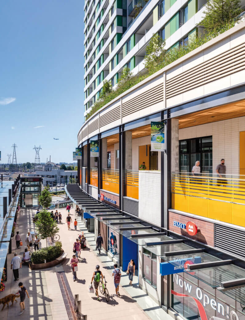 A bustling urban plaza with shops and pedestrians, featuring greenery and a residential building in the background under a blue sky.