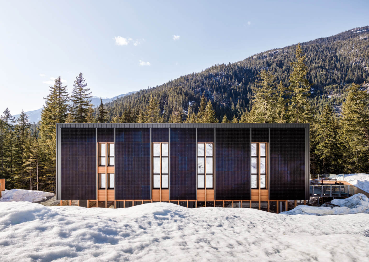 Sleek building design with wooden accents, surrounded by snow and trees, set against a mountainous backdrop under a clear sky.