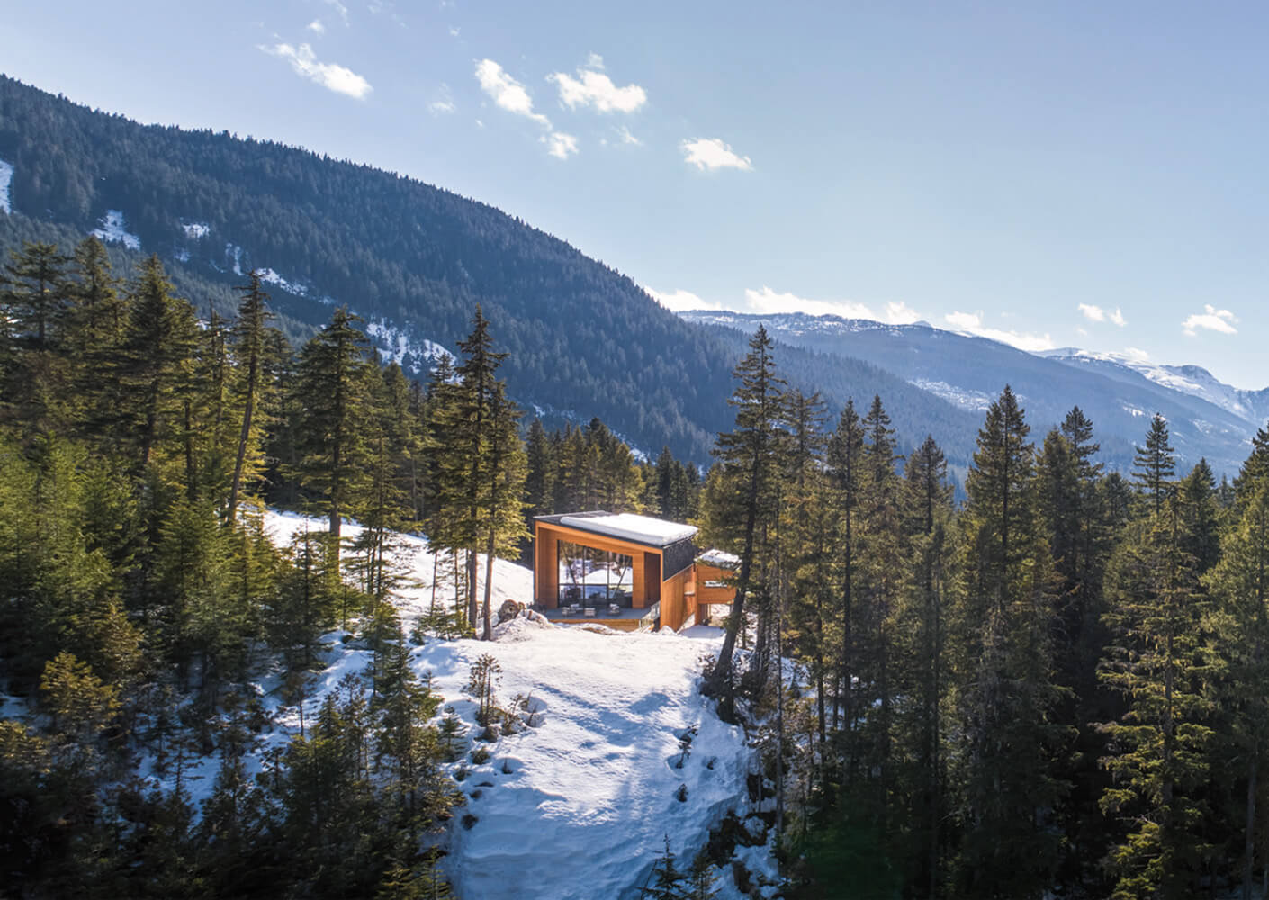 Aerial view of a cabin surrounded by snow and forested mountains, featuring a wooden exterior and large windows. Clear blue sky in the background.