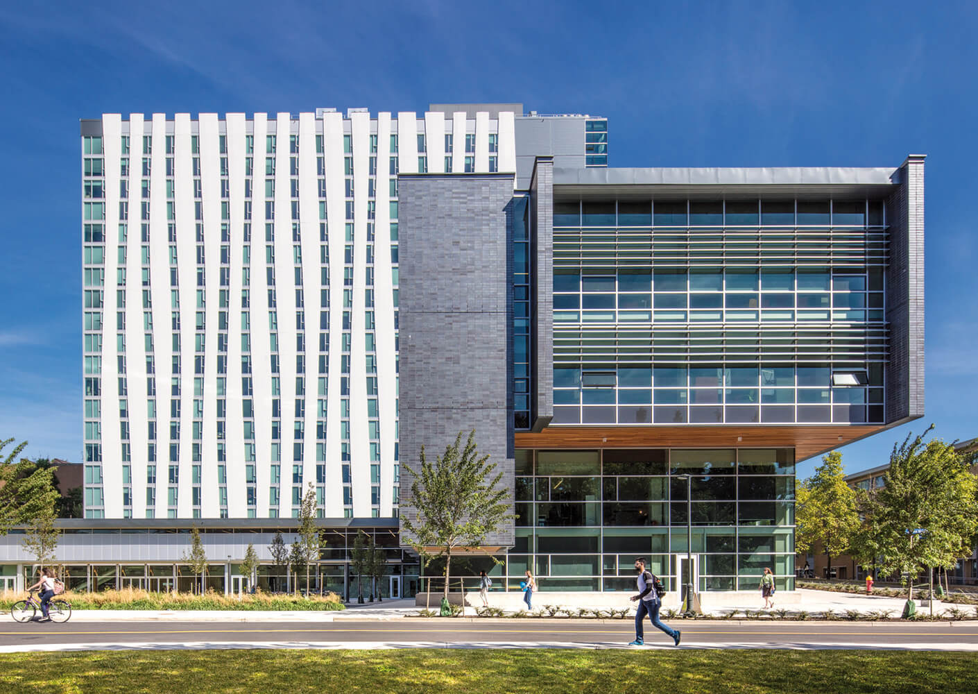 University building featuring glass and stone elements, with people walking and biking in front of landscaped grounds on a sunny day.