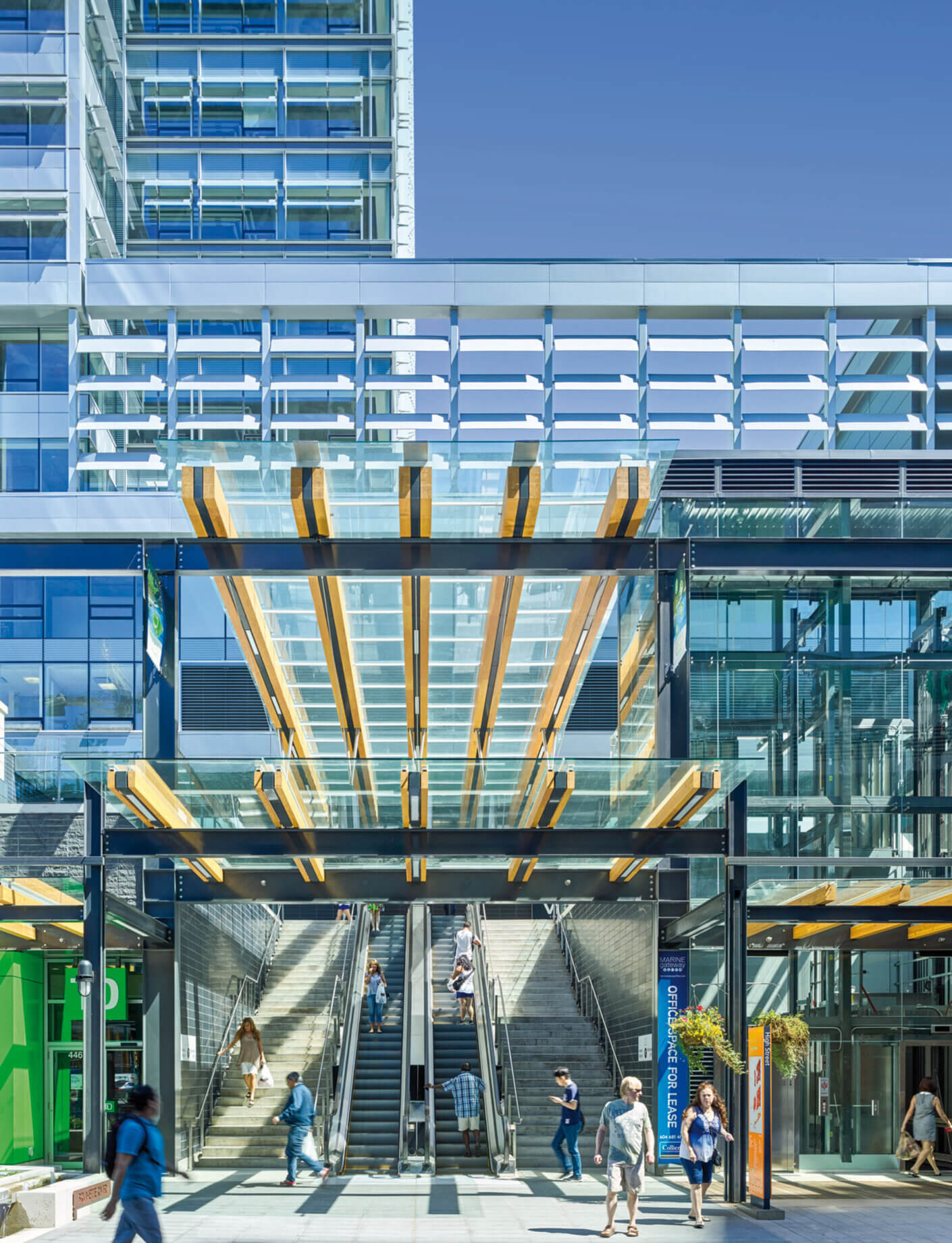 Urban commercial space featuring glass and wood elements, with people using escalators and stairs in a bustling environment.