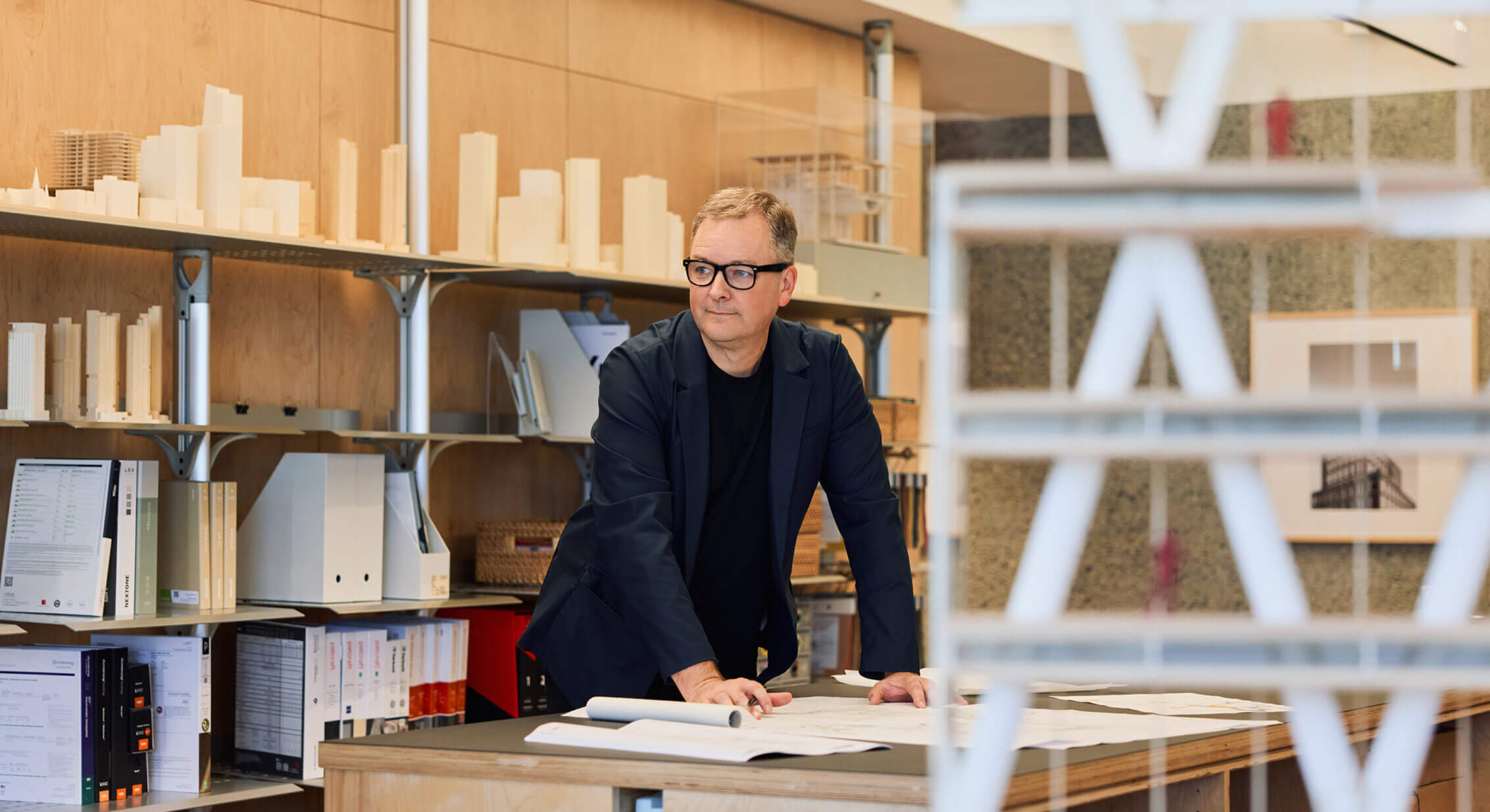 Architect in an office with architectural models and design materials, reviewing plans on a desk, surrounded by organized shelving.