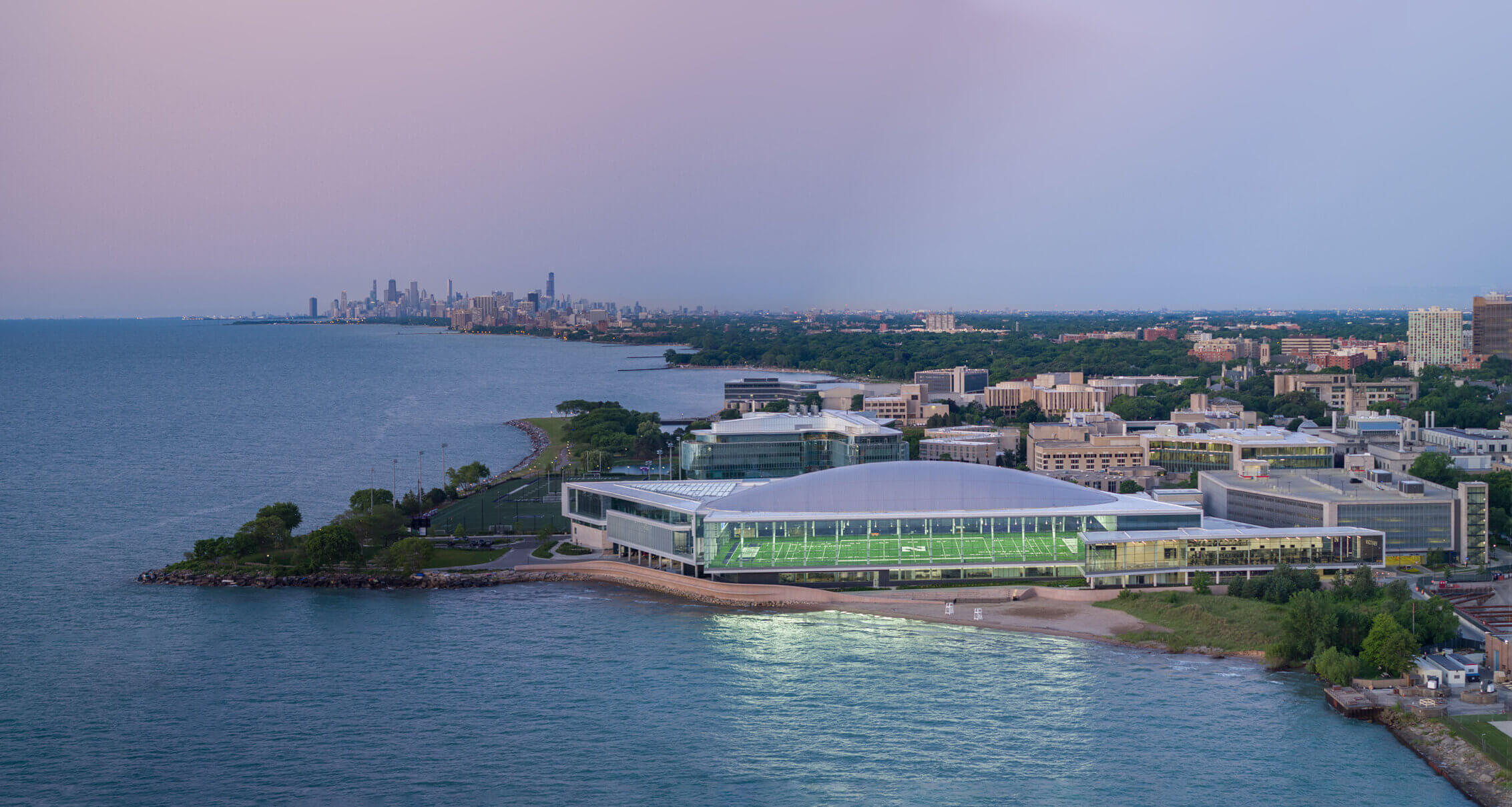 Aerial view of a sports and events center along the shore, with a city skyline in the background and a lake in the foreground.