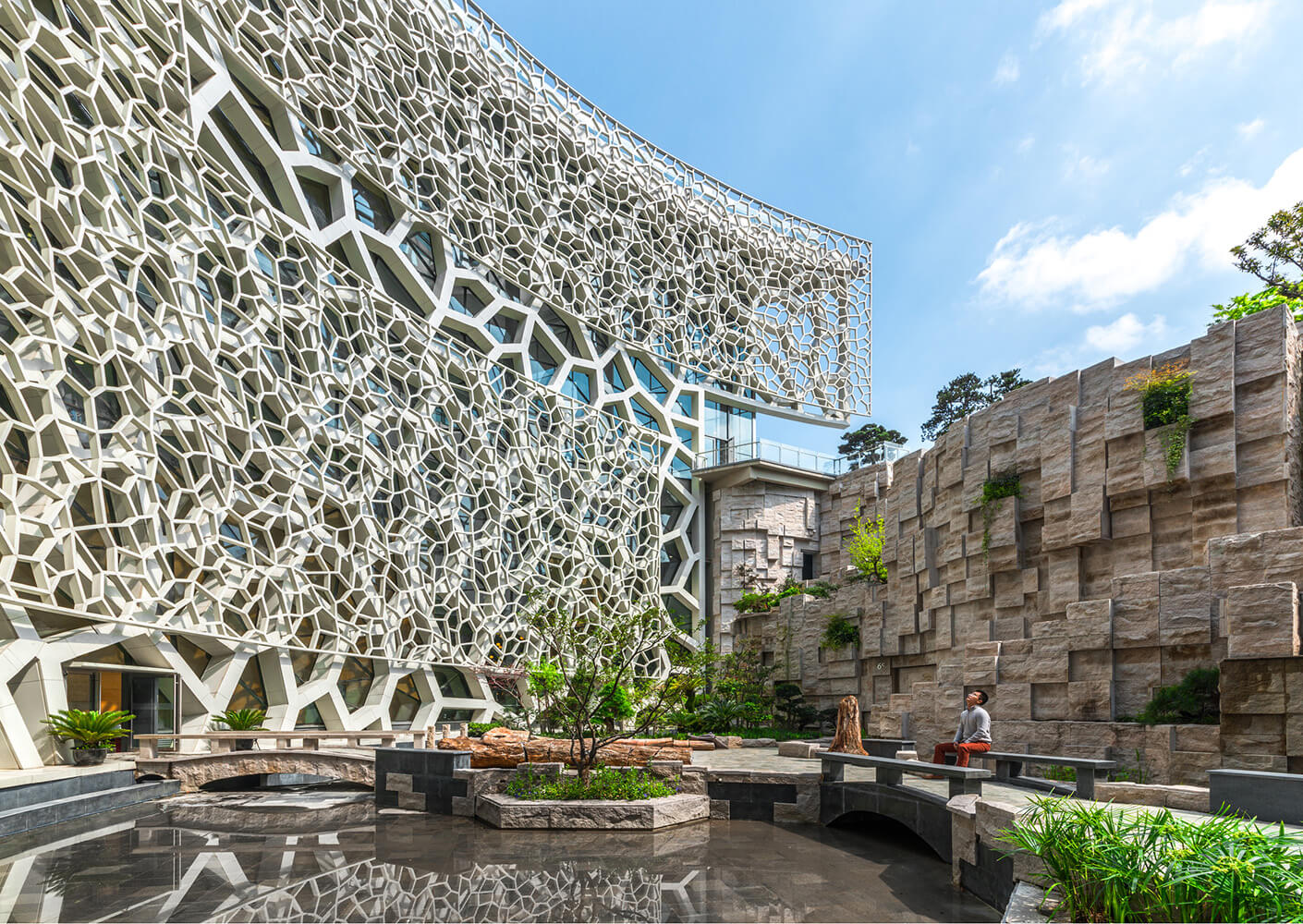 A unique architectural structure featuring intricate patterns, surrounding greenery, and a tranquil water feature in an outdoor space.