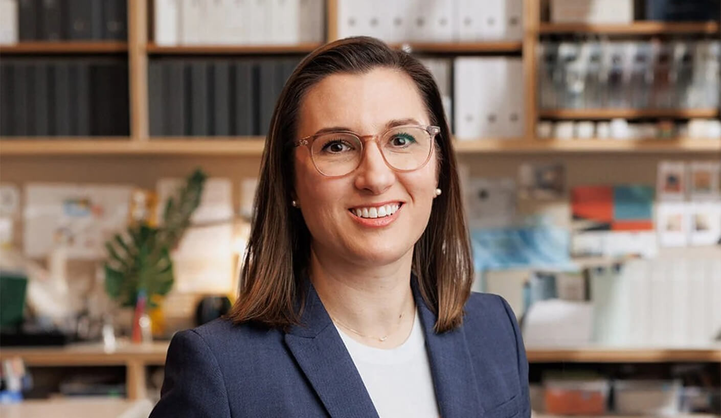 Portrait of a woman smiling in an office setting, wearing glasses and a blazer, with shelves of binders and design materials in the background.