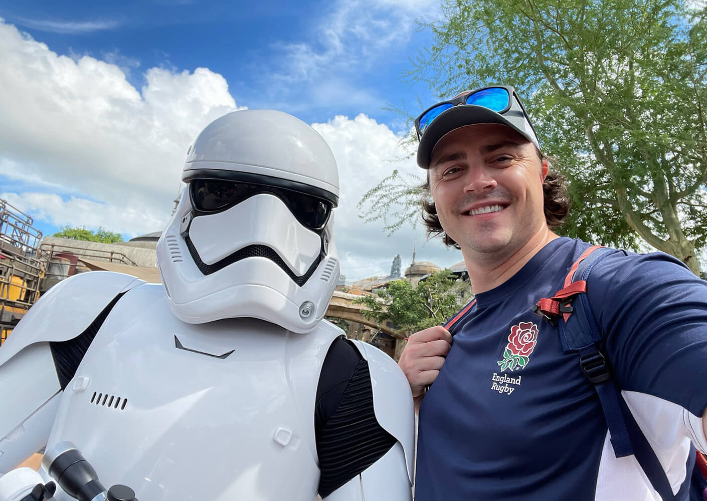 A man smiles for a selfie with a Stormtrooper at a theme park, showcasing Star Wars-themed attractions under a blue sky.