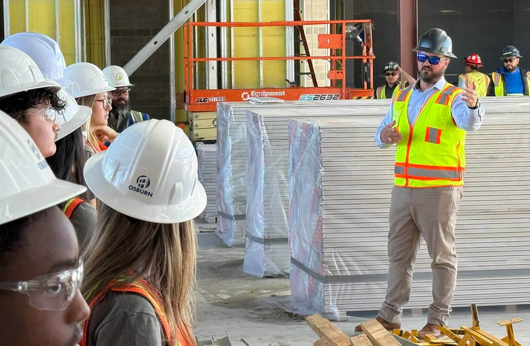 Danny leading a student tour of a construction site, wearing a hard hat, safety vest, and safety goggles.