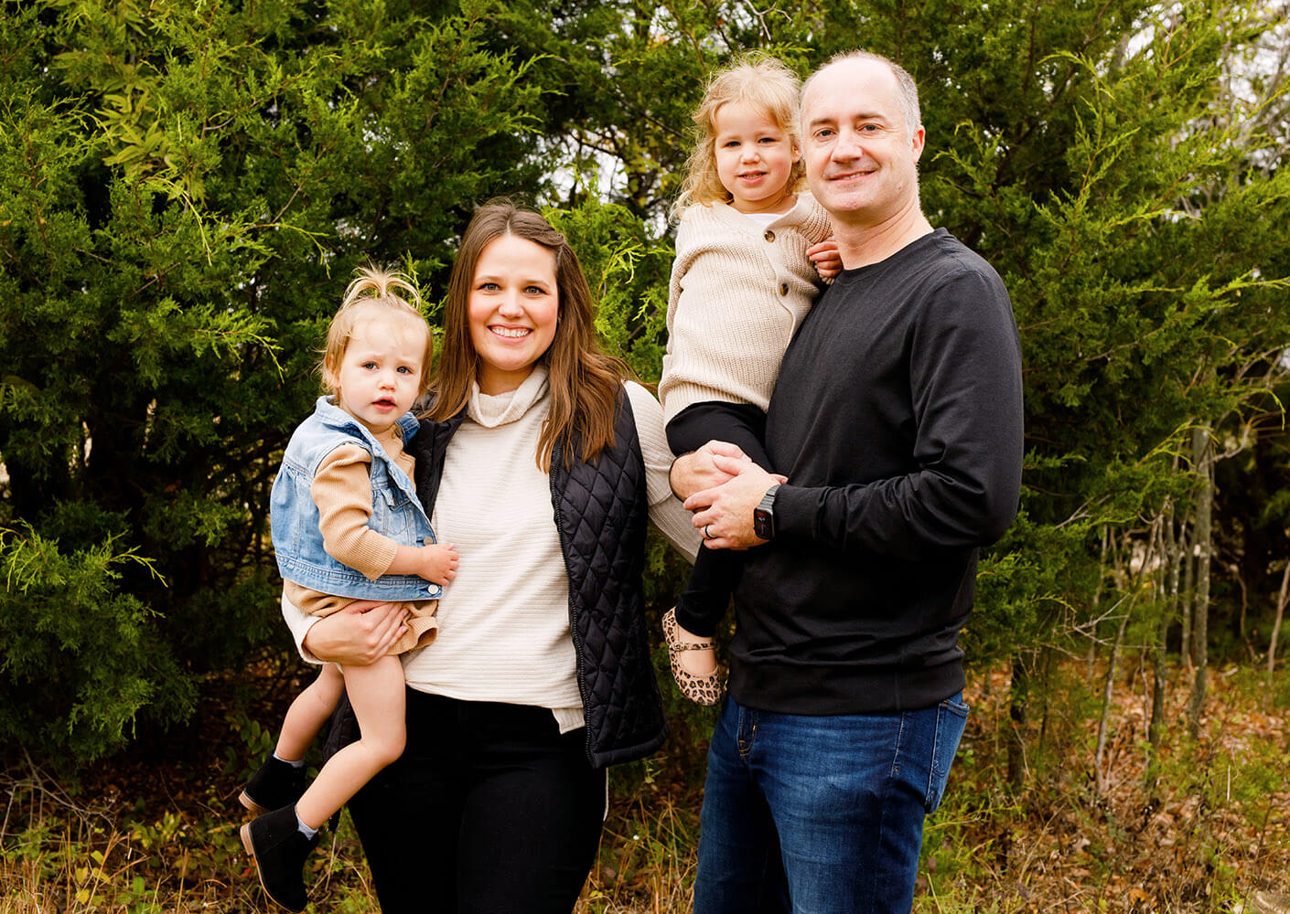 Lauren, husband Kevin Mereness, and their two daughters pose for family photo set against a background of greenery.