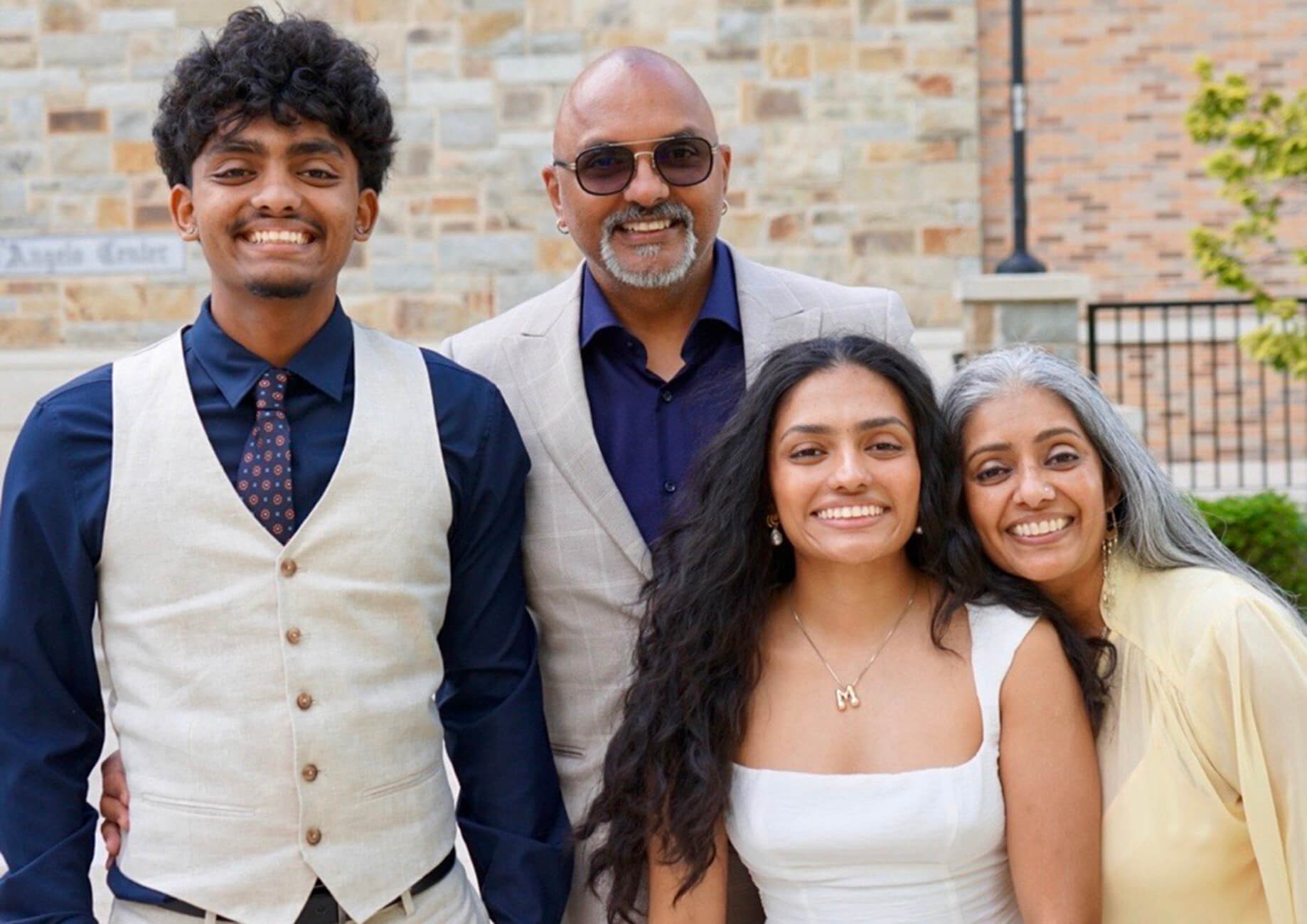 Nirmala smiles with her husband, son, and daughter outdoors. They wear formal attire against a stone building backdrop.