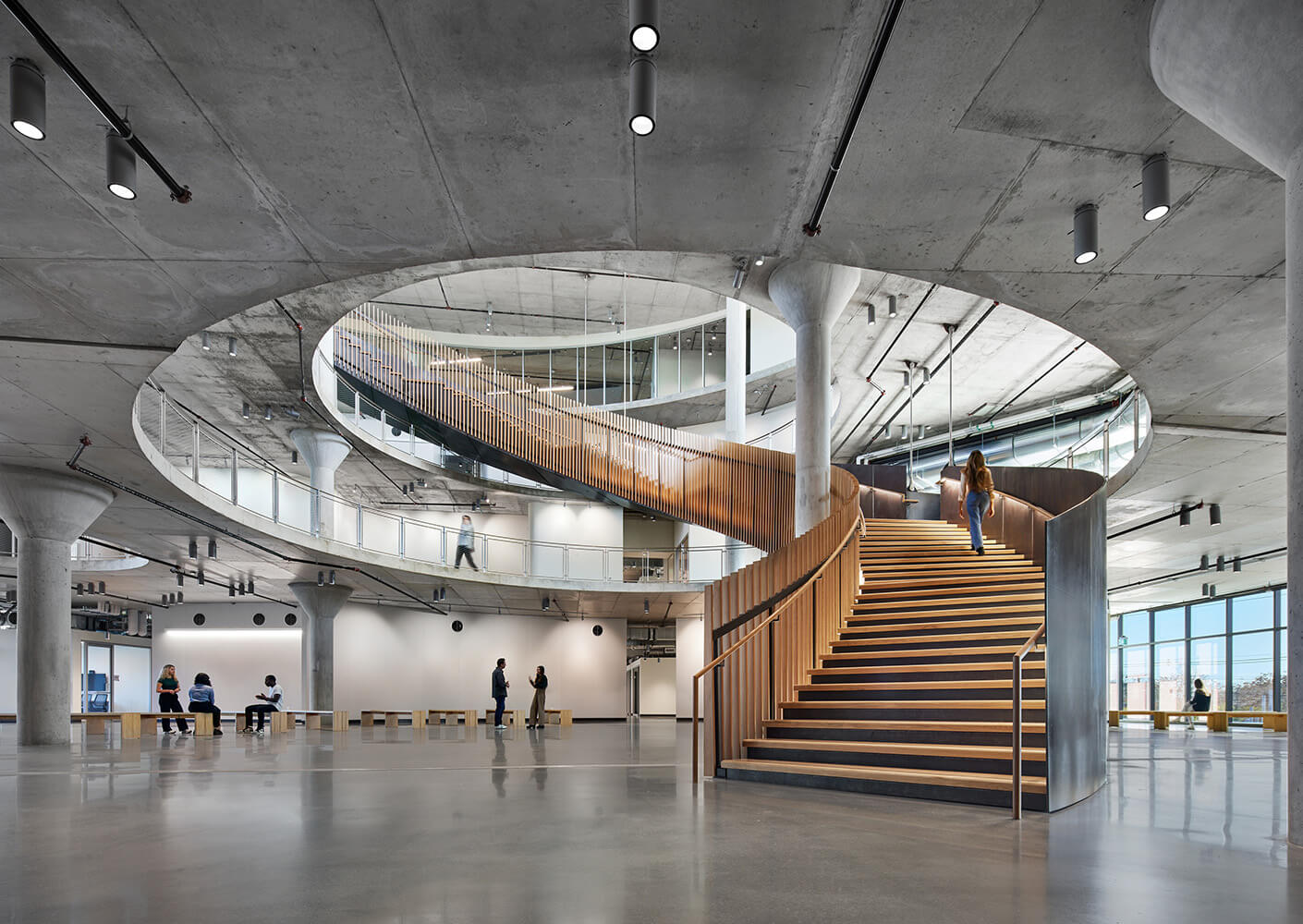 Interior view of a spacious architecture featuring a prominent wooden staircase, concrete pillars, and people engaging in various activities.