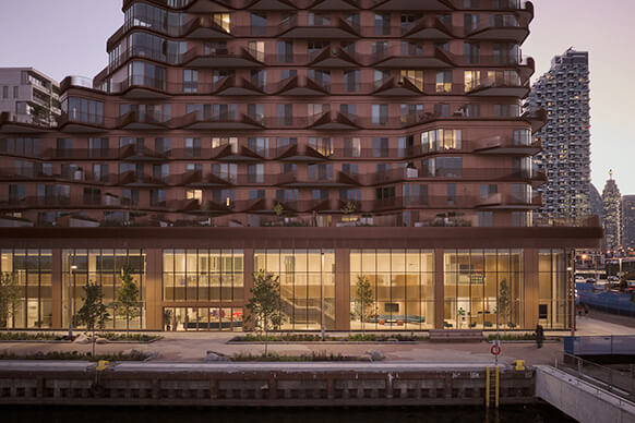 Architectural view of a building with a wave-like façade, featuring large glass windows and greenery along the waterfront promenade.