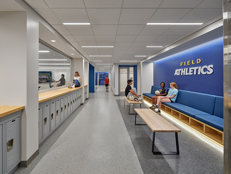 Athletic facility interior featuring a corridor with lockers, seating areas, and signage. Students engage in activities and relax.