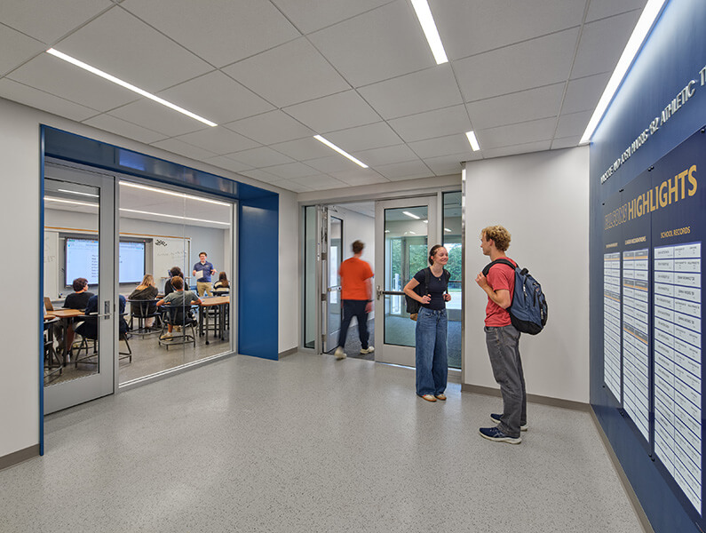 Two students converse in a hallway with large glass doors leading to a classroom. A whiteboard is visible in the background.