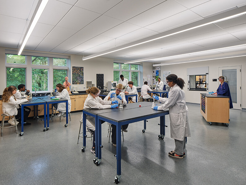 Students in lab coats work collaboratively in a science lab, engaging in experiments and discussions at various tables under bright lighting.