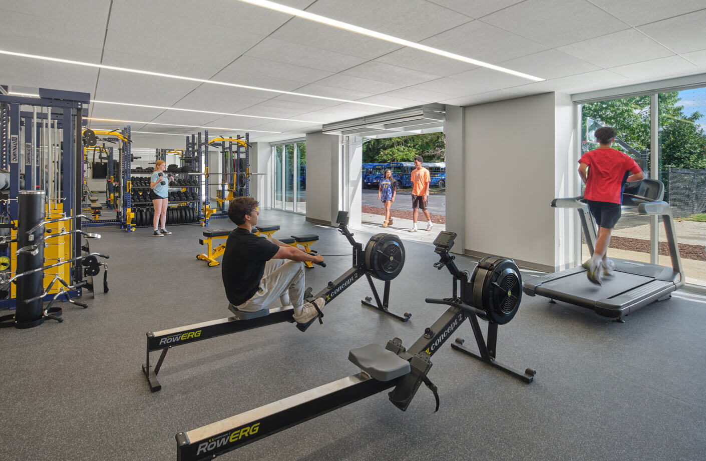 Fitness center interior featuring workout equipment, including rowing machines and a treadmill, with individuals exercising and socializing.