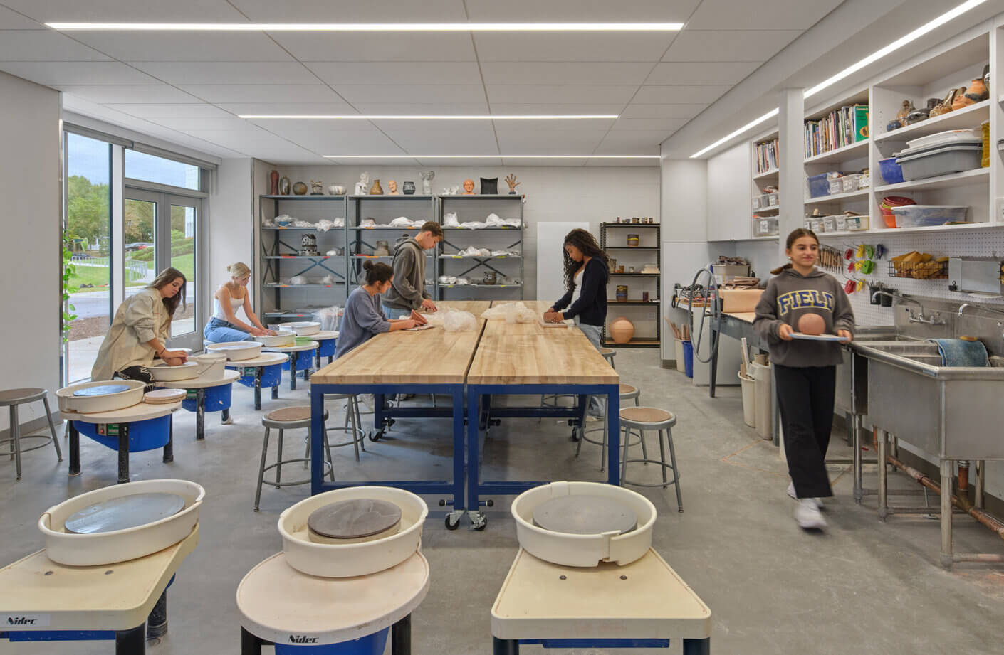 Students engaged in pottery making in a well-lit studio with pottery wheels, worktables, and shelves of ceramic pieces.