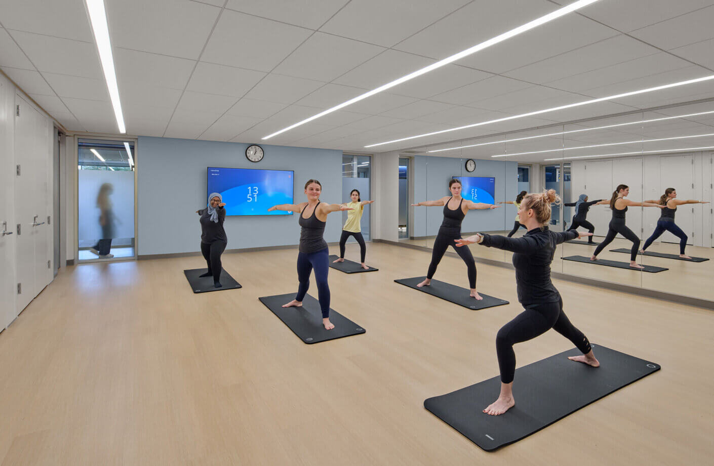 A yoga class in a spacious studio with participants practicing poses on mats, mirrors reflecting their movements, and a digital clock on the wall.