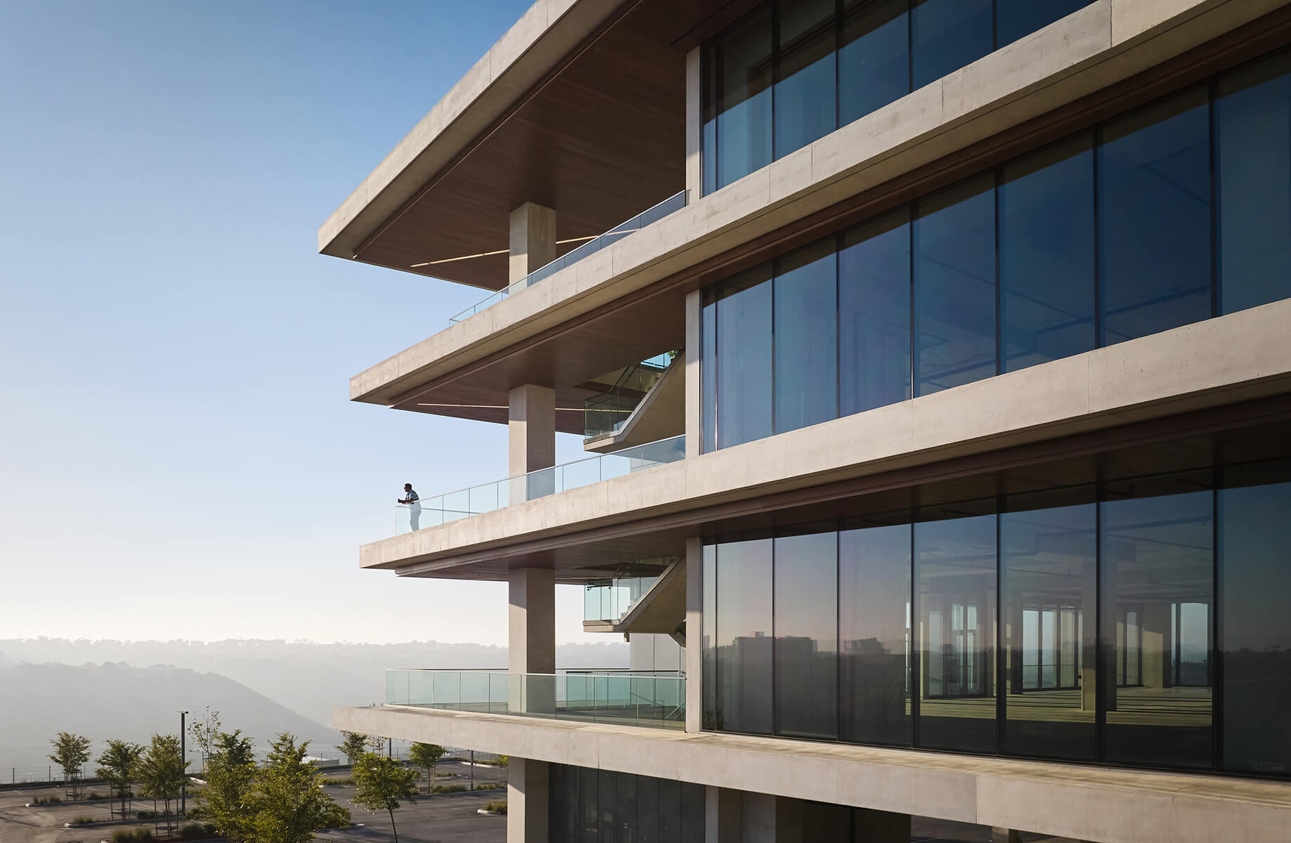 A contemporary building with large glass windows, showcasing multiple floors and a balcony with a person overlooking a scenic view.