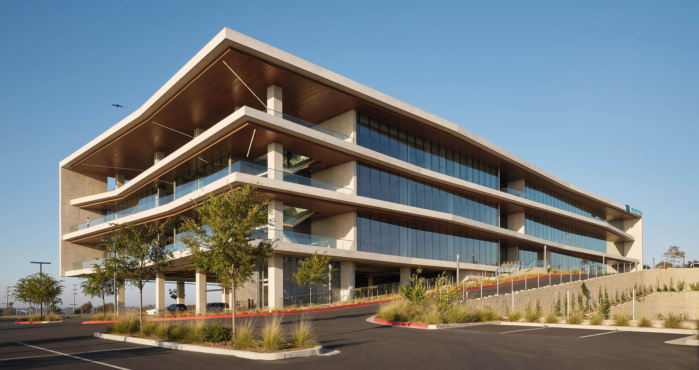 Contemporary office building with large glass windows, tiered balconies, and landscaped surroundings, set in a parking lot against a blue sky.