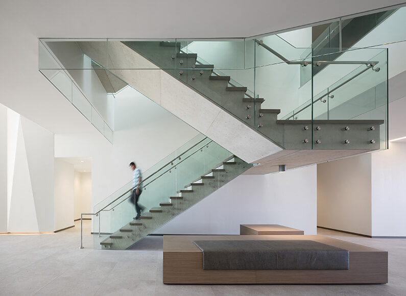 Interior staircase with glass railings and concrete steps; a person descends the stairs in a spacious, well-lit environment.