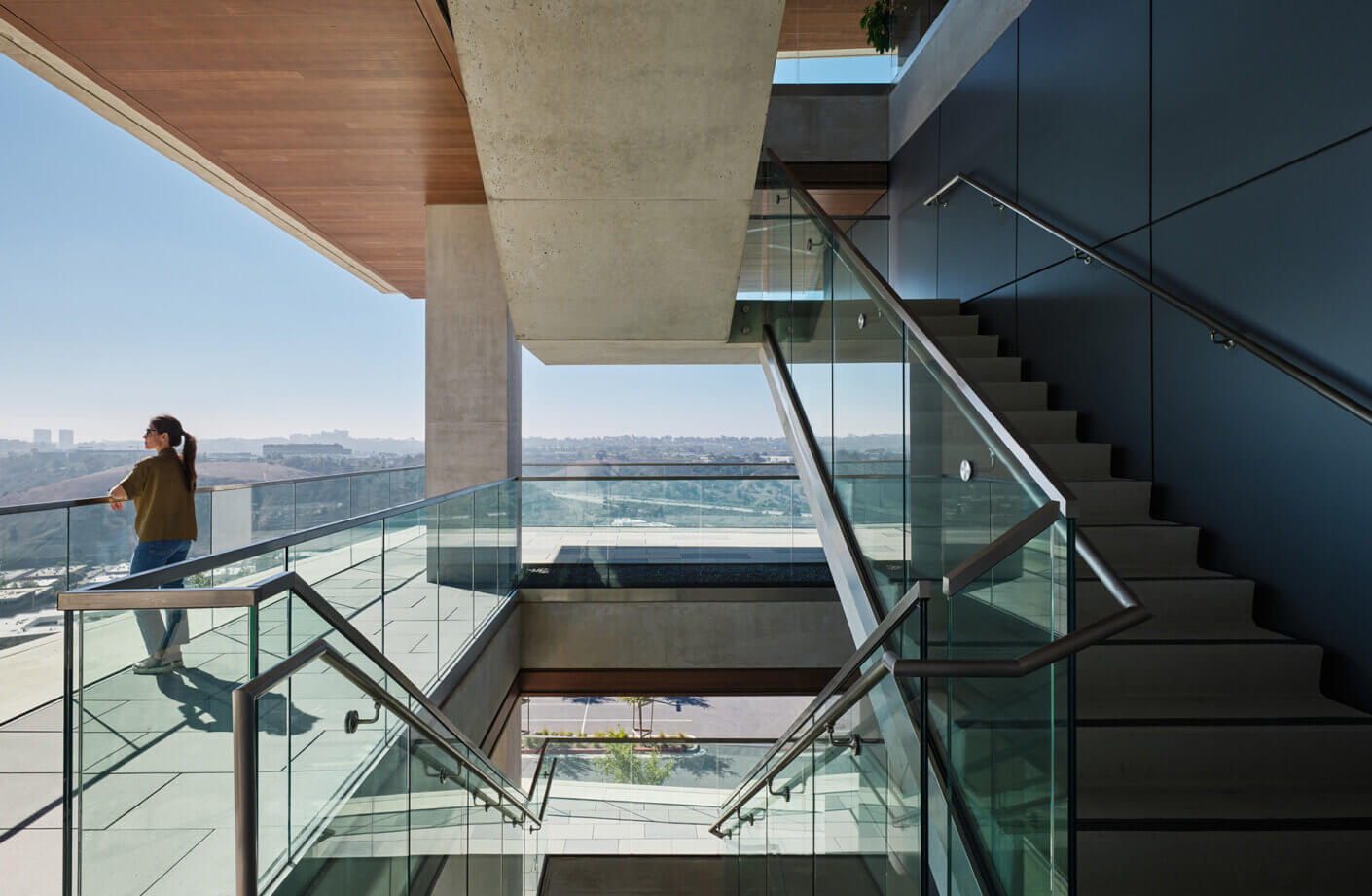 Woman standing on a glass balcony overlooking a cityscape, next to a concrete stairwell with modern handrails and ample natural light.