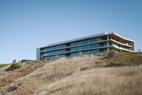 A hillside building with large glass windows, featuring multiple levels and a flat roof, surrounded by dry grass and sparse vegetation.