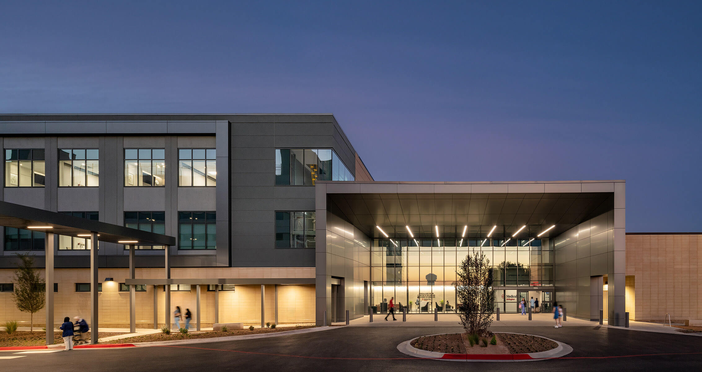 New healthcare facility entrance at dusk, featuring large glass windows and a sleek architectural design. Pedestrians approach the entrance.
