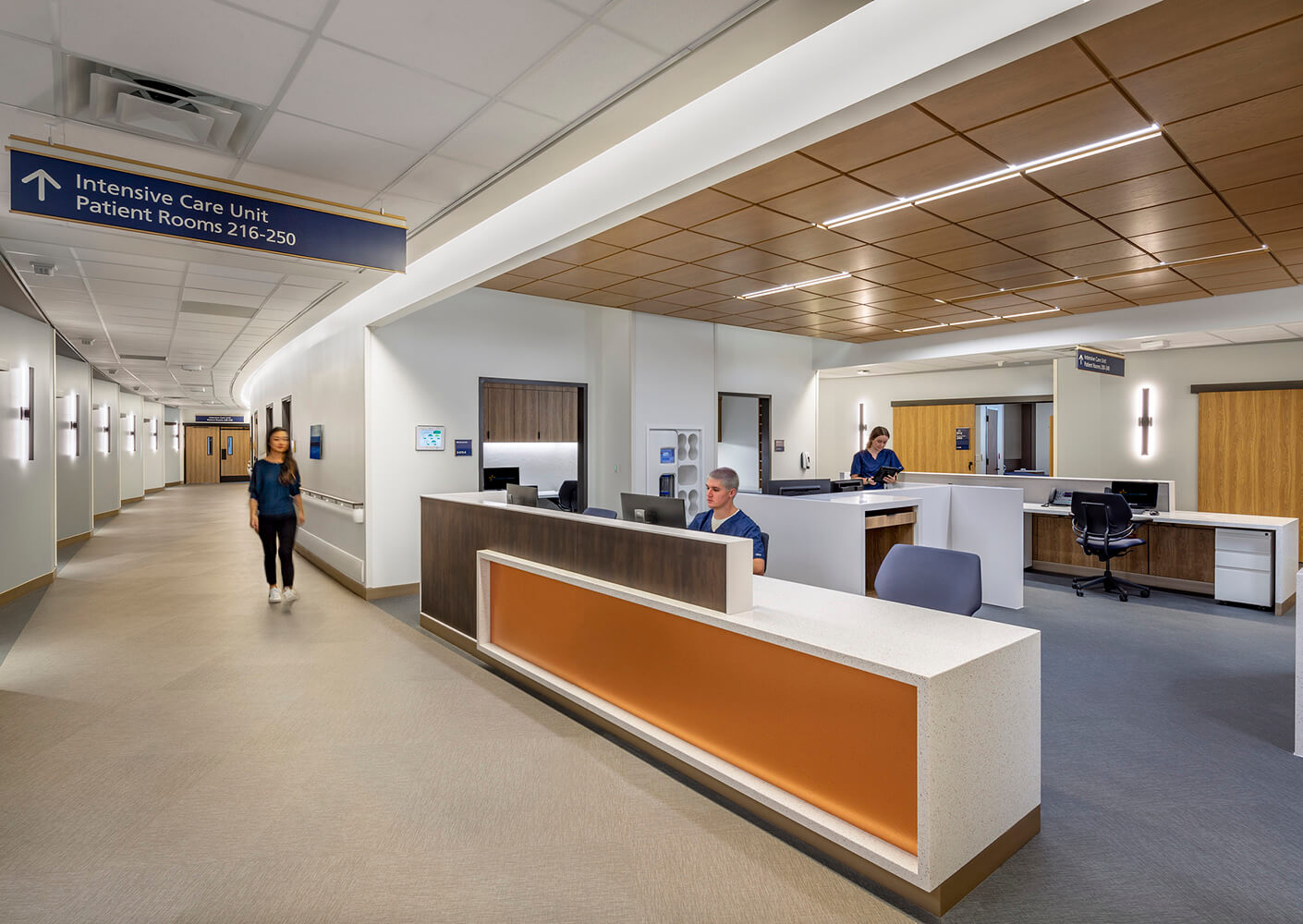 Hallway of an intensive care unit featuring staff at a reception desk, directional signage, and adjacent patient rooms.