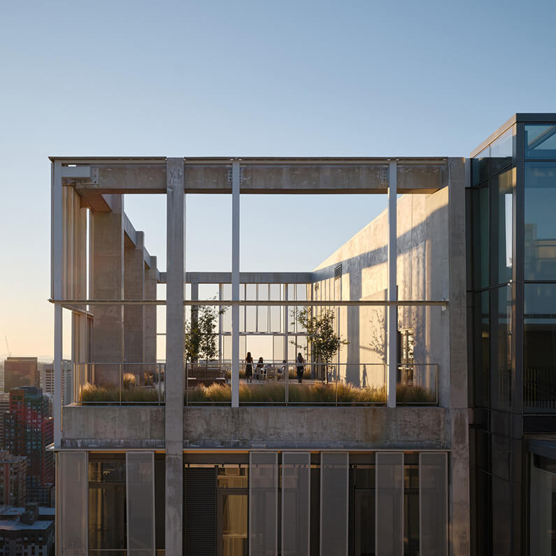 Rooftop terrace with seating and greenery on a concrete and glass building, featuring expansive views of the city skyline at sunset.