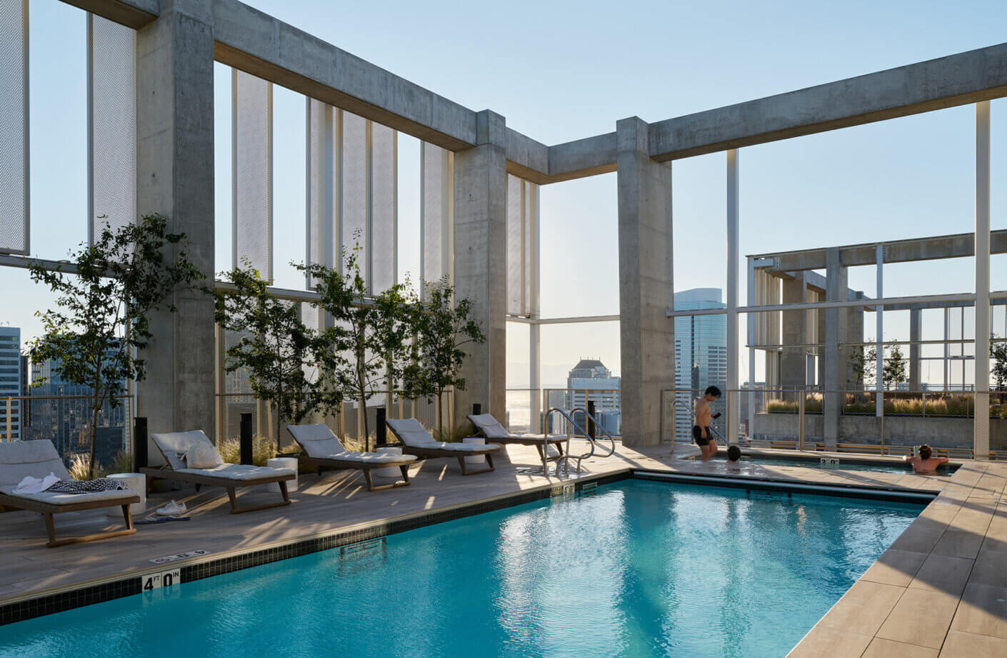 Rooftop pool area featuring sun loungers, greenery, and city skyline views. Two individuals engage in fitness activities near the water.
