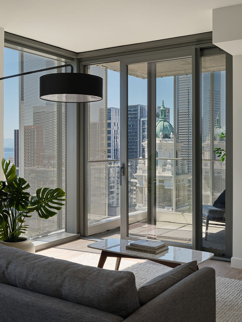 Interior view of a living space featuring a sofa, coffee table, potted plant, large windows with cityscape, and modern light fixture.