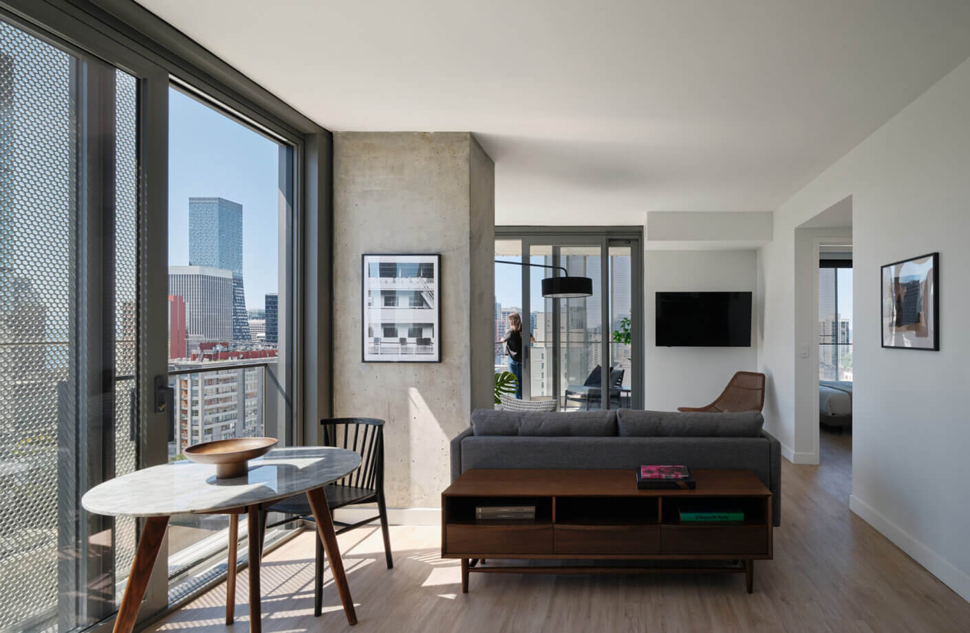 Interior view of a stylish apartment featuring a gray sofa, round table, and large windows with cityscape views. Natural light fills the space.