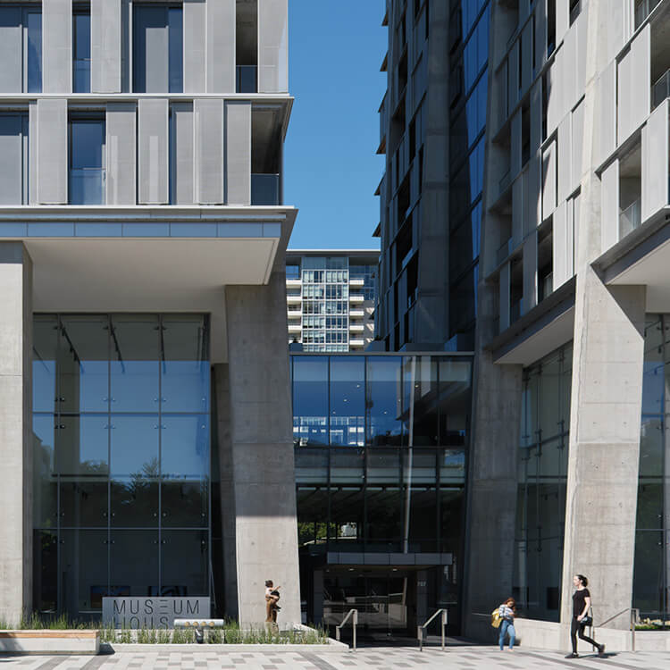 Entrance of a building with large glass windows and a prominent sign reading MUSEUM HOUSE, surrounded by concrete structure and modern urban design elements.