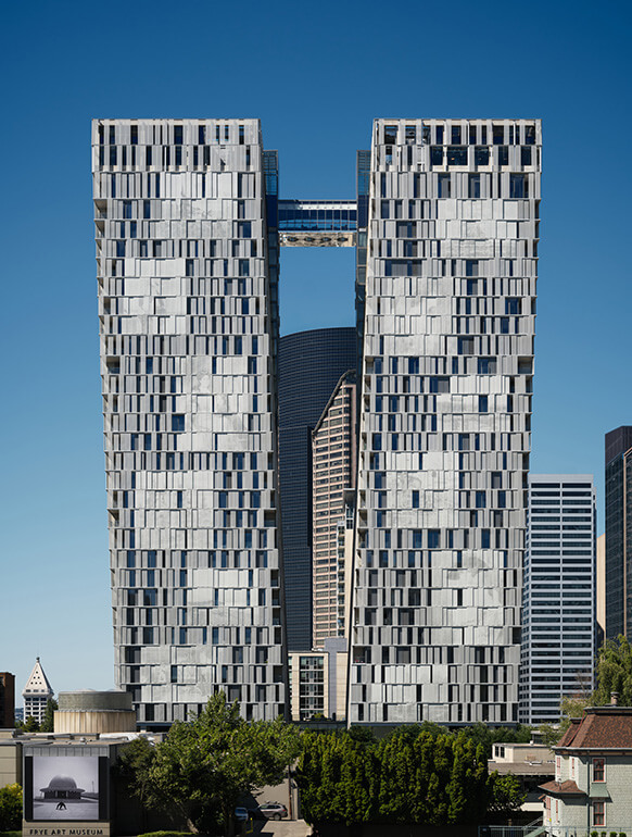 High-rise building with an innovative design, featuring two towers connected by a bridge, surrounded by urban landscape and clear blue sky.