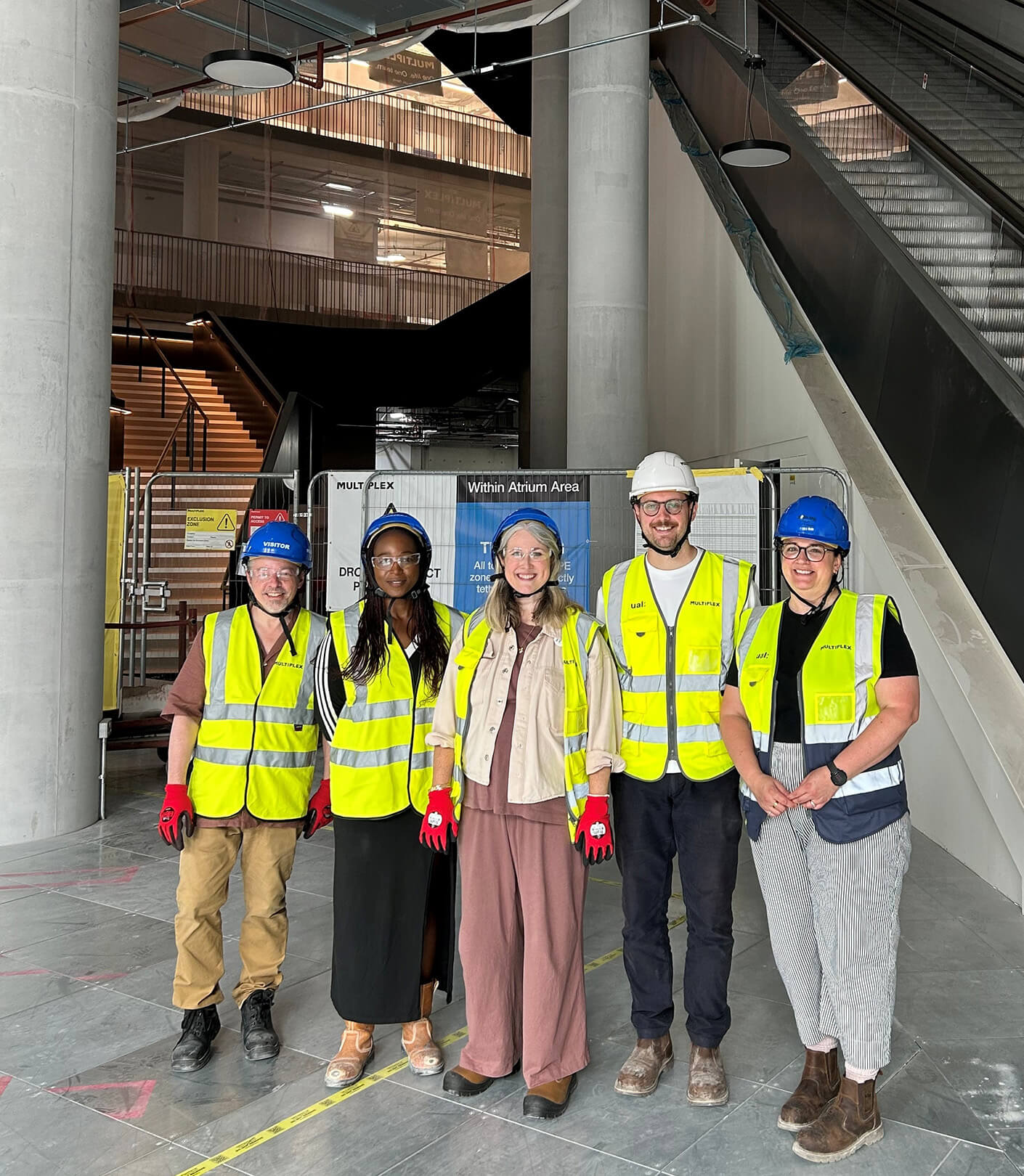 Five construction workers in safety gear pose for a photo inside a building under construction, featuring scaffolding and warning signs.