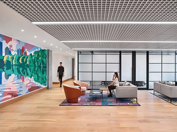 Office interior featuring seating areas, a vibrant mural, and large windows. Two people engage in the space, highlighting a collaborative atmosphere.