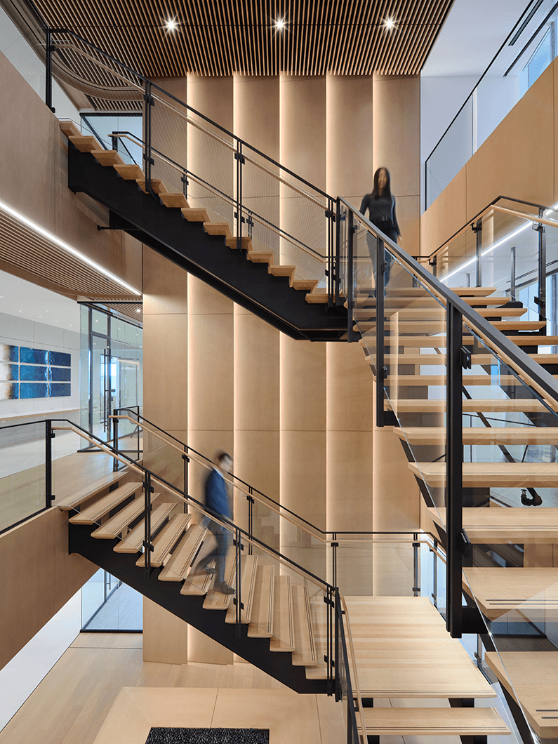 Stairs with wooden treads and metal railing in a well-lit interior space. Two people walk down and up the staircase.