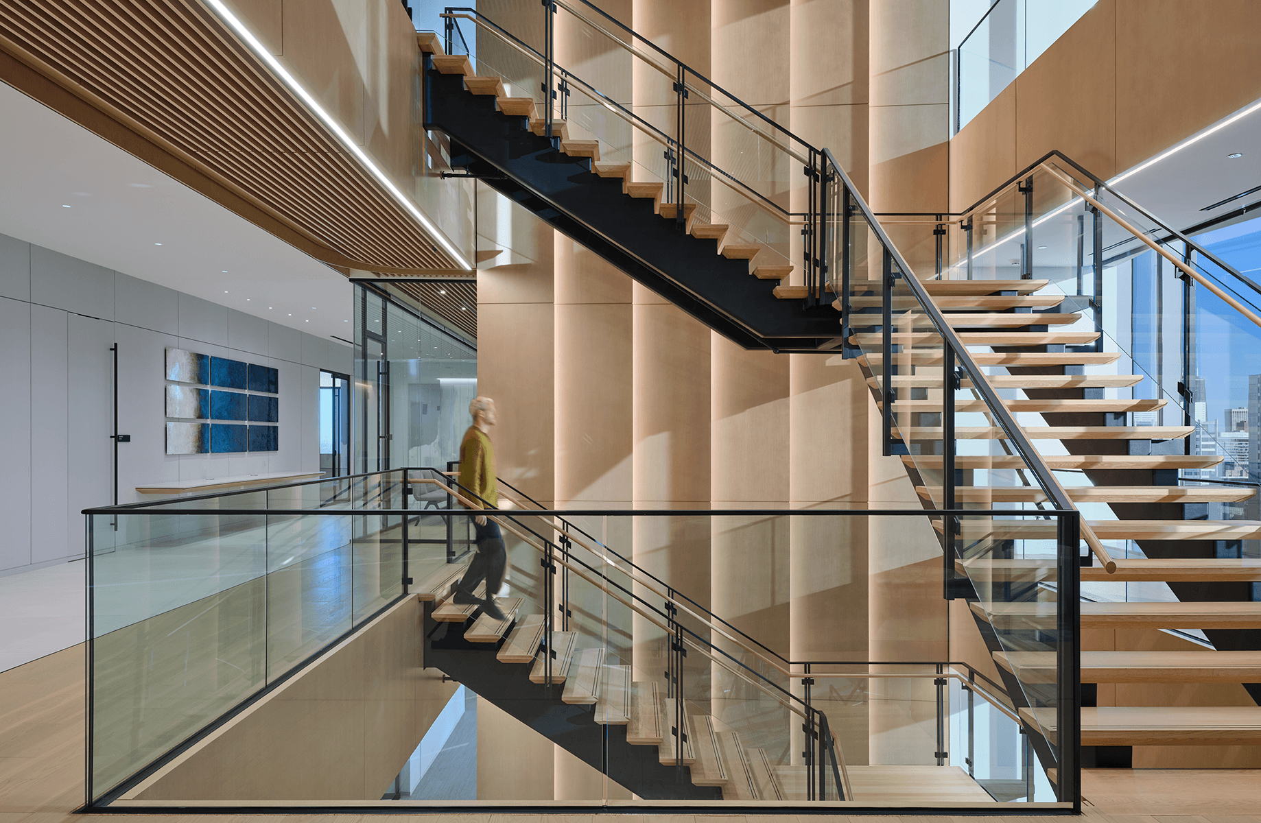Interior view of a multi-level staircase with wood steps and glass railings. A person ascends the stairs in a well-lit space.