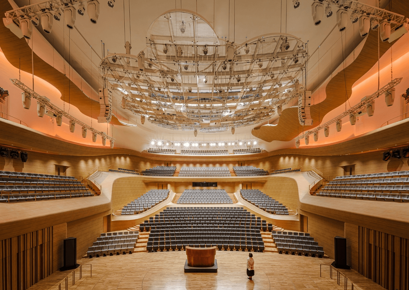 Interior view of a concert hall featuring tiered seating, wooden panels, and suspended lighting fixtures, with a small figure on stage.