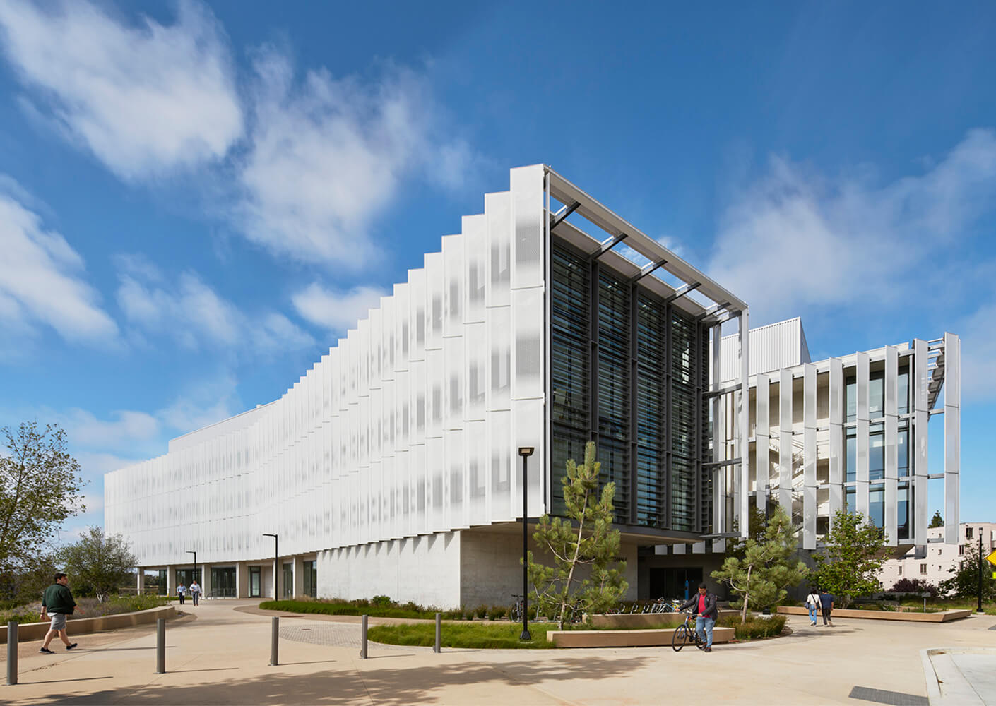 Exterior view of a building featuring a unique façade, landscaped pathways, and people walking and biking on a clear day.