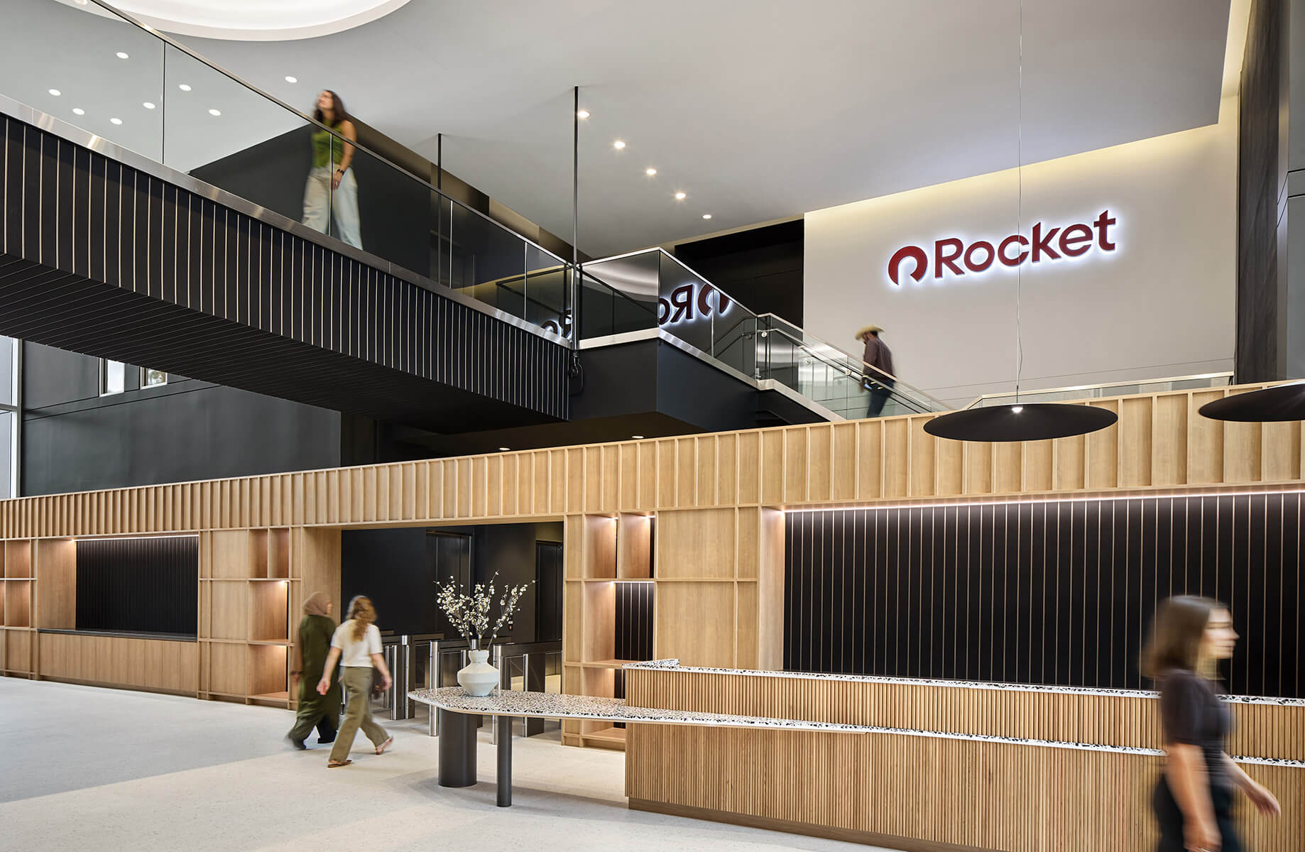 Office reception area featuring a wooden desk, seating, and a balcony above. People walk through the space with a visible Rocket logo.