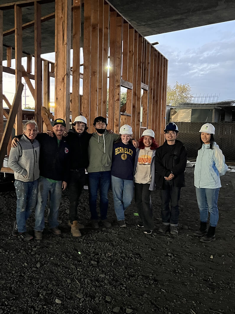 Group of eight people in hard hats posing for a photo at a construction site with wooden framing in the background during sunset.