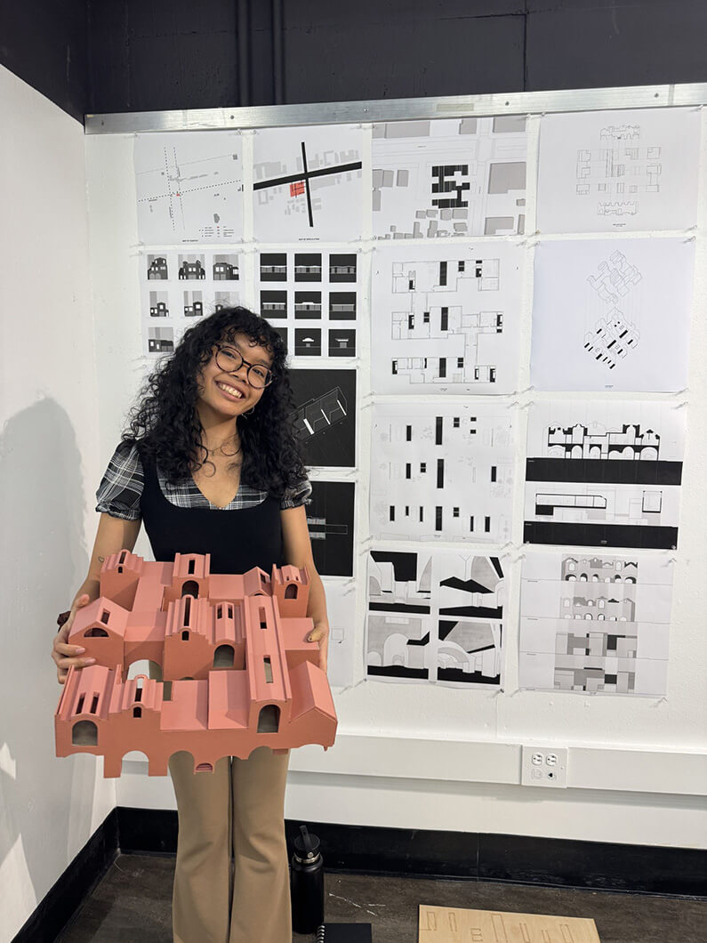 Student holding a pink architectural model in front of design sketches on a wall, showcasing a blend of textures and layouts.