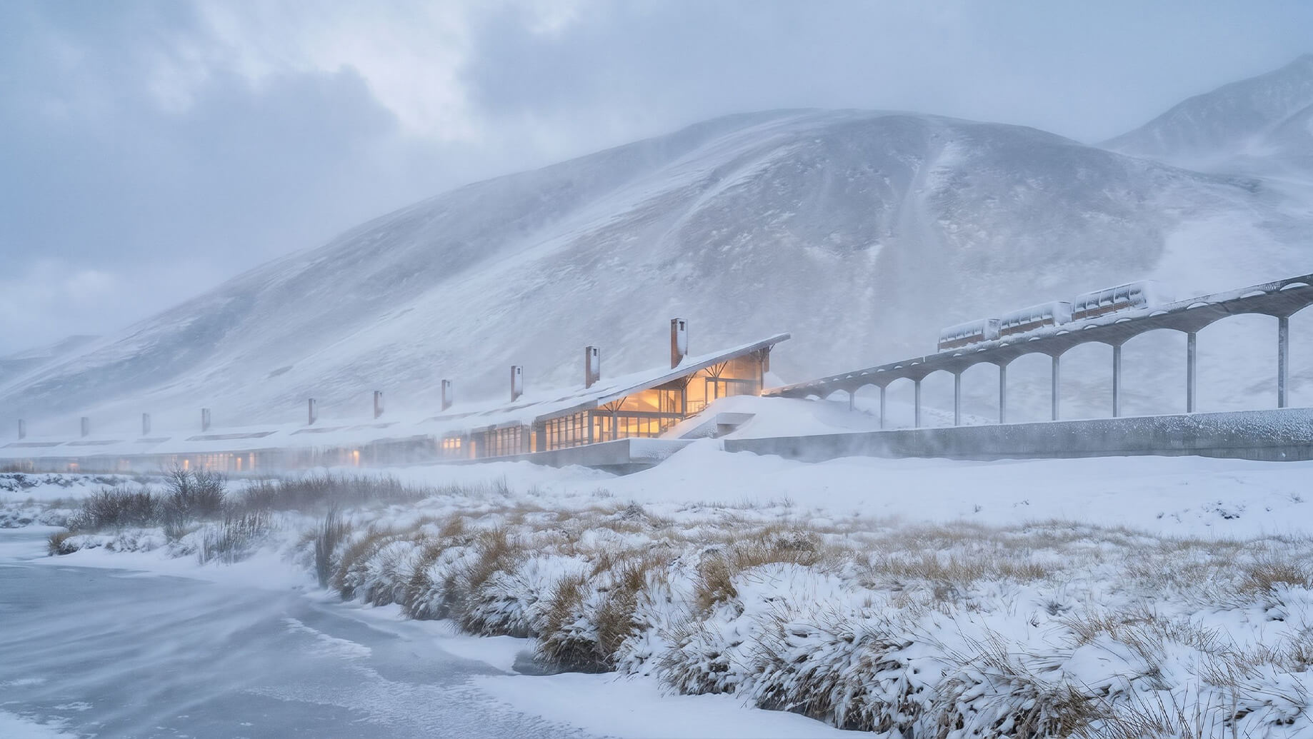 Snow-covered landscape featuring a building with large windows, adjacent to a train track, surrounded by mountains and heavy snowfall.