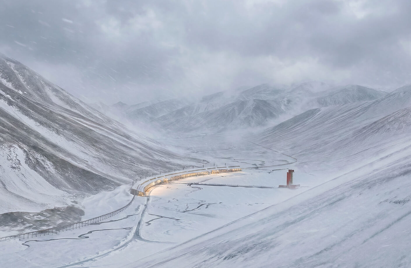 Snow-covered mountains with a winding road and illuminated buildings in a remote landscape under a cloudy sky during a snowstorm.